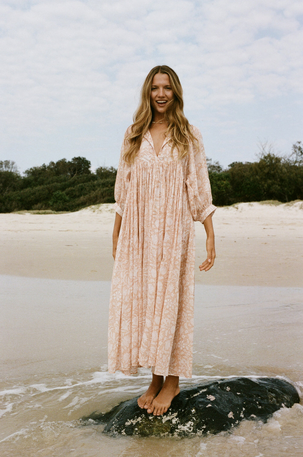 Daughters of India Kyra Maxi Dress in Apricot Blush — standing on rock in surf smiling, sand dunes behind