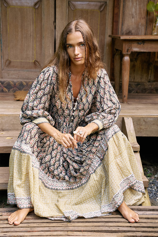 Bhoomi Maxi Dress Onyx — full length front view seated on wooden steps, onyx hand block print maxi dress with contrast tiered border hem, rustic wooden doors and furniture | Daughters of India