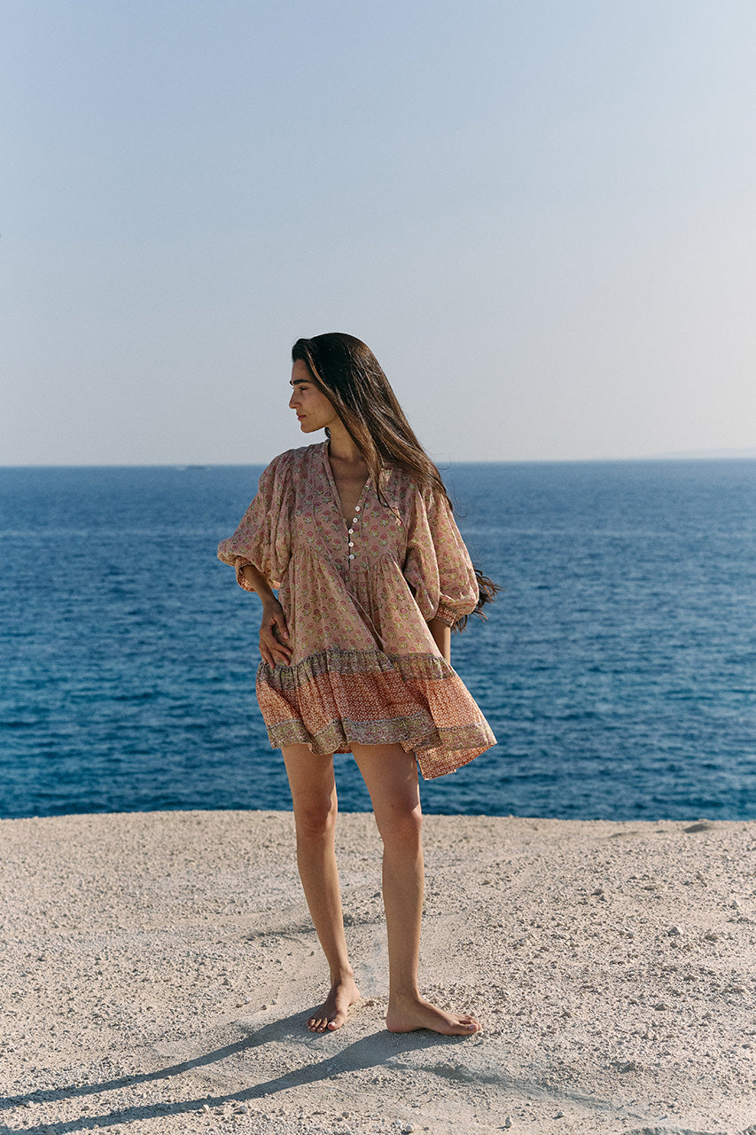 Woman standing on a white ledge by the ocean wearing the Daughters of India Bhoomi Mini Dress in Sunset, the peach floral mini dress in relaxed flowing silhouette