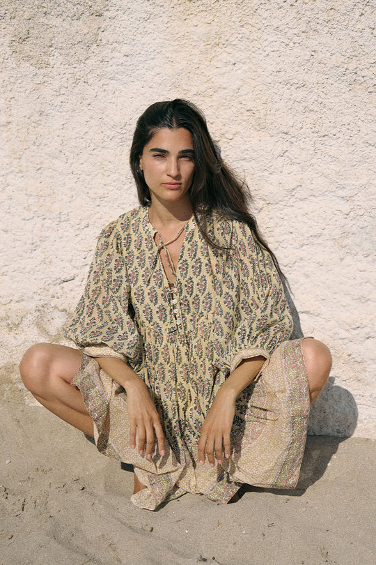 Woman sitting cross-legged on sand wearing the Daughters of India Bhoomi Mini Dress in Meadow, a sage green and cream paisley block print cotton mini dress with billowed sleeves