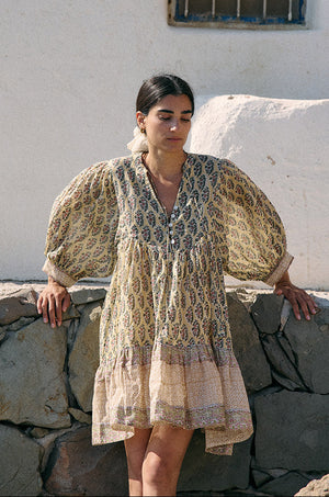 Woman leaning against a stone wall wearing the Daughters of India Bhoomi Mini Dress in Meadow, showing the large billowed sleeves and tiered hem with border print detail