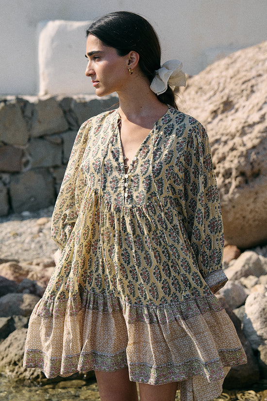 Woman with a white flower in her hair beside a stone wall by water wearing the Daughters of India Bhoomi Mini Dress in Meadow, showing the V-neckline and tiered skirt hem