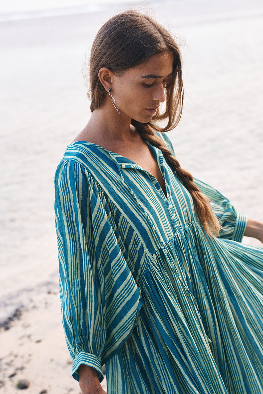 Woman with a braided hairstyle at the beach wearing the Daughters of India Kyra Midi Dress in Spearmint, the green striped fabric flowing in the sea breeze