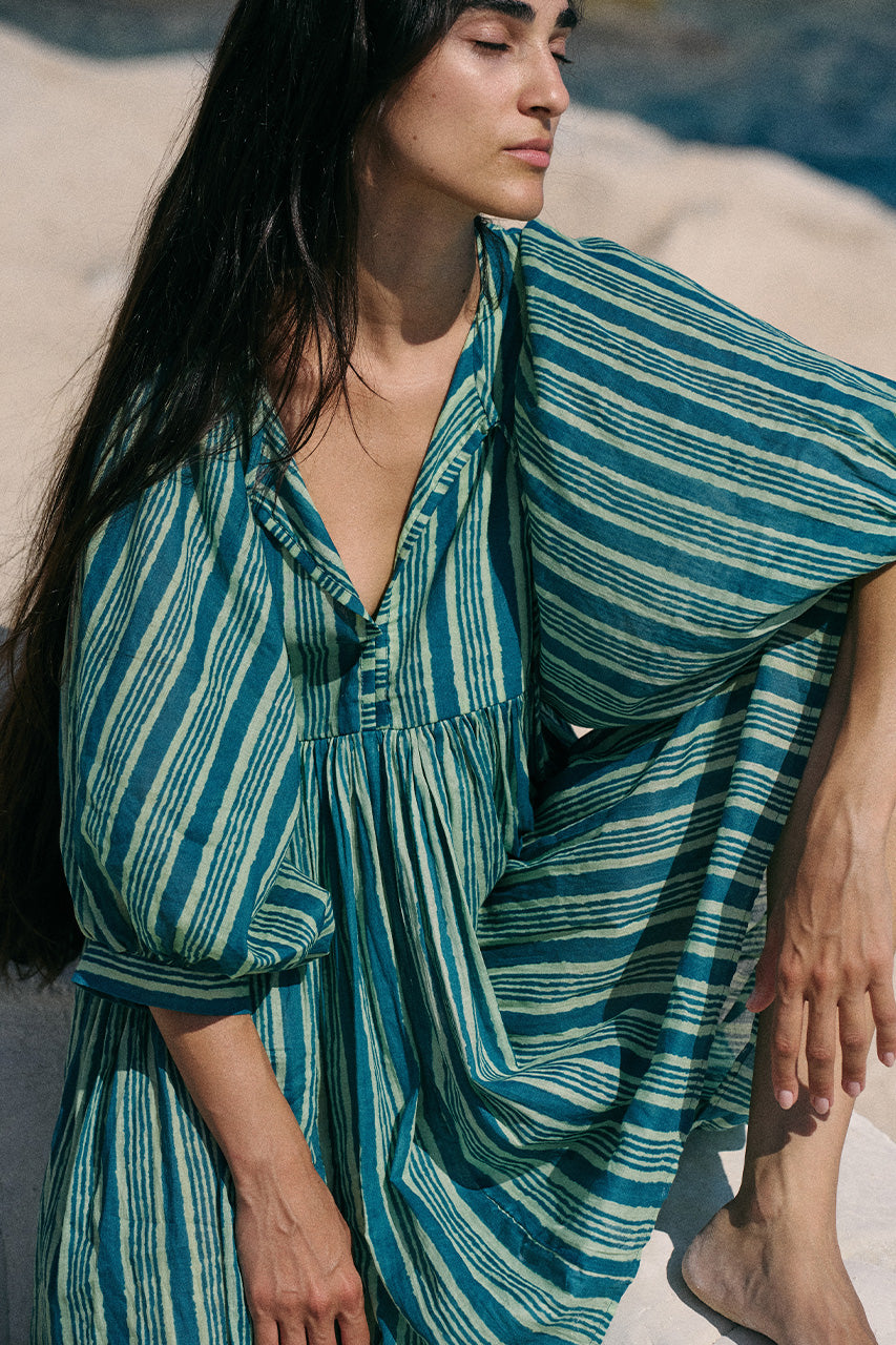 Woman wearing a green and blue striped dress sitting on a sandy beach.
