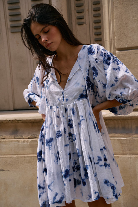 Kyra Mini Dress Deep Sea — close-up front view at stone building entrance, deep sea shibori tie-dye cotton mini dress with V-neckline and balloon sleeves, European shuttered facade | Daughters of India