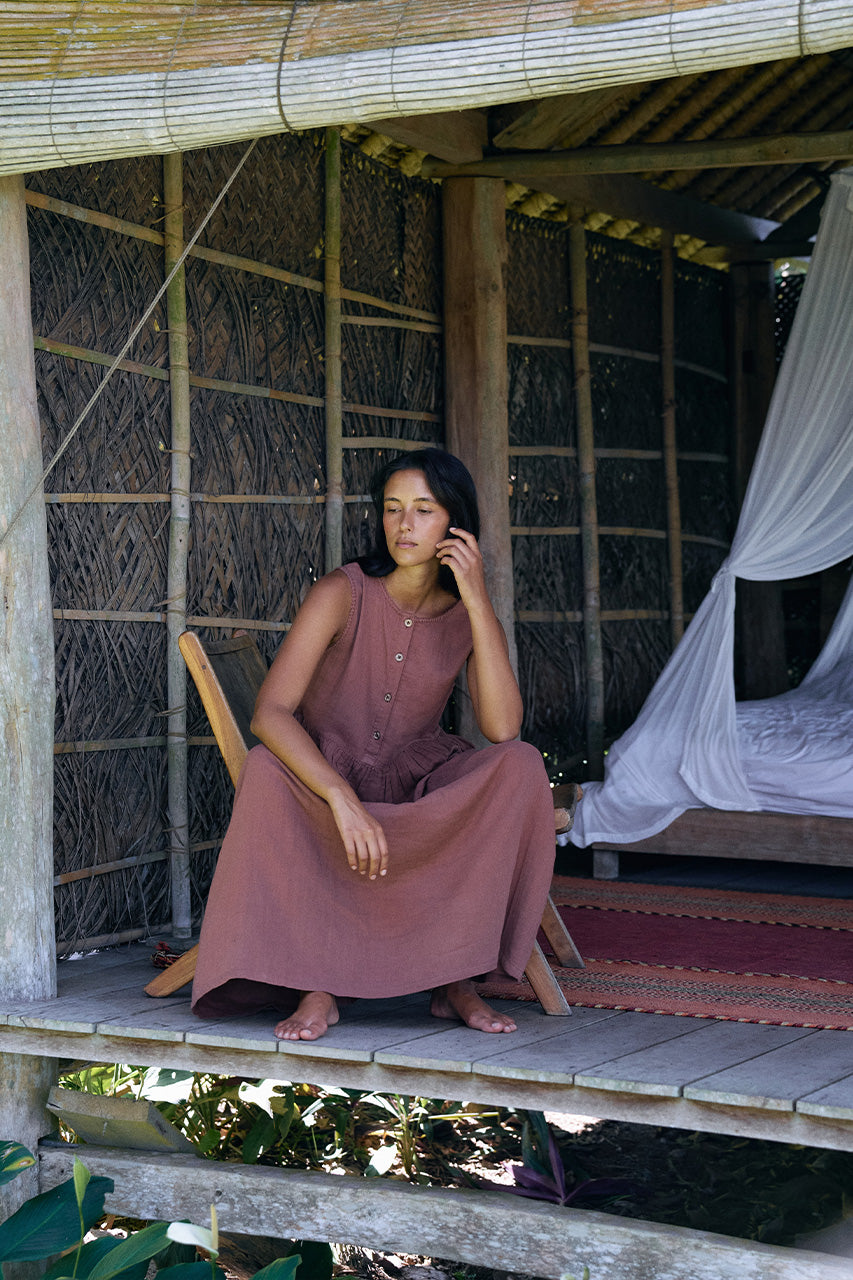 Nadi Dress Chocolate — seated in wooden chair on elevated bamboo hut veranda, chocolate brown double gauze cotton midi dress with wooden button front, woven palm walls and white mosquito net | Daughters of India